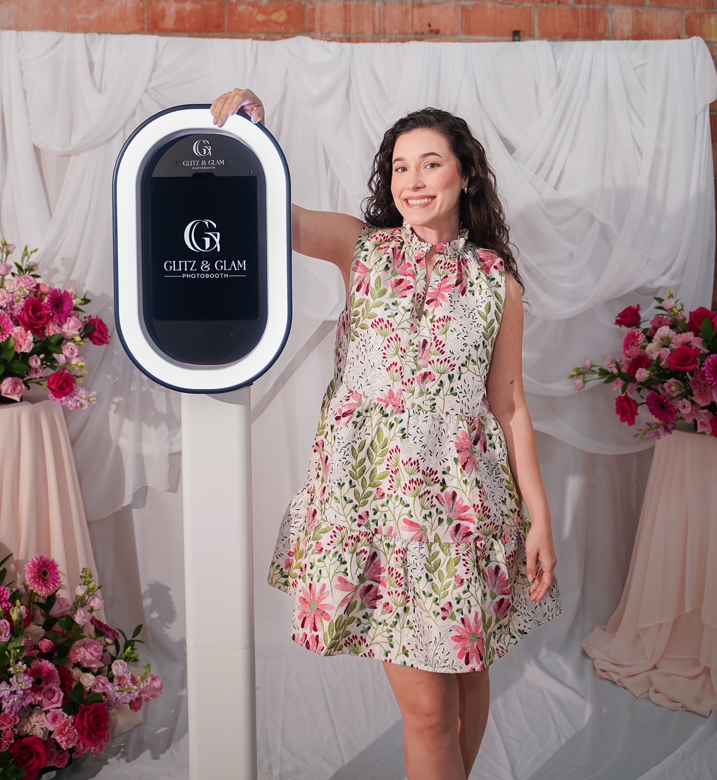 A smiling woman beside the Glitz & Glam photo booth, framed by white draping and pink flowers